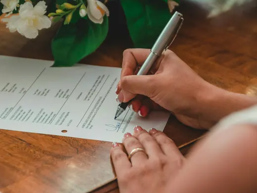 Une main qui signe un document sur une table de bois, près d’un bouquet de fleurs blanches