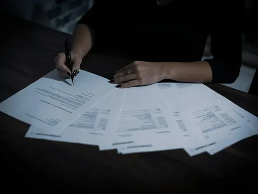 Des feuilles étalées sur une table sombre, une main tient un stylo